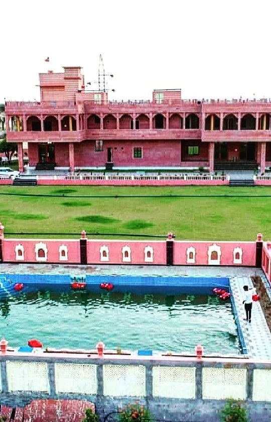 Aerial daytime view of Sri Karni Heritage Resort showing sandstone building, huge green lawn and swimming pool